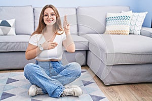 Young caucasian woman sitting on the floor at the living room smiling swearing with hand on chest and fingers up, making a loyalty