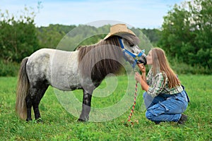 Young Caucasian woman is sitting on the grass and kissing her horse in its muzzle, side view
