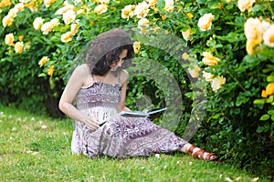 Young Caucasian woman sitting on green grass near yellow roses bush in a rose garden, reading a book, looking to the book