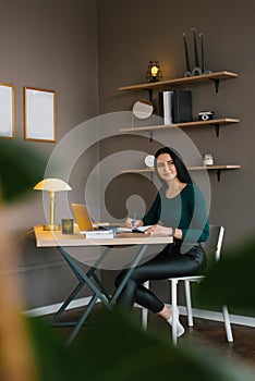 Young Caucasian woman is sitting at home at her desk with a laptop and writing something in a notebook