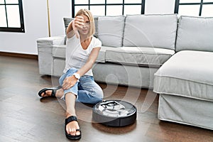 Young caucasian woman sitting at home by vacuum robot looking unhappy and angry showing rejection and negative with thumbs down