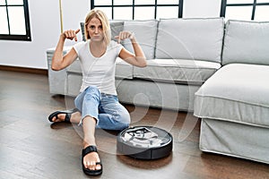 Young caucasian woman sitting at home by vacuum robot pointing down looking sad and upset, indicating direction with fingers,