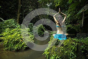 Young Caucasian woman sitting on the rock, practicing yoga and pranayama. Raising arms with namaste mudra. View from back. Self