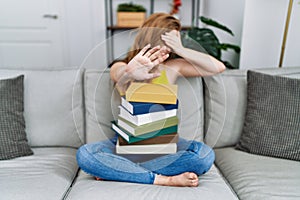 Young caucasian woman sitting on the sofa with books at home covering eyes with hands and doing stop gesture with sad and fear