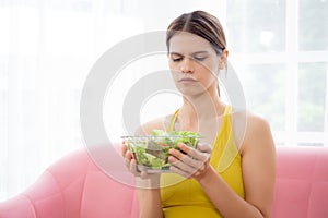 Young caucasian woman sitting on sofa holding vegetable salad while feeling dislike and expression disgusted.