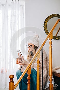 Young caucasian woman sitting on a stairs at home, drinking tea and using mobile phone