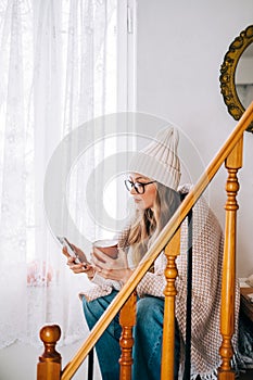 Young caucasian woman sitting on a stairs at home, drinking tea and using mobile phone