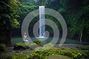 Young Caucasian woman sitting on the stone near the waterfall in Vajrasana or Diamond pose. Hands in namaste mudra. Tropical