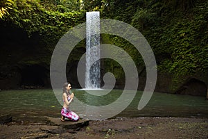 Young Caucasian woman sitting on the stone near the waterfall in Vajrasana or Diamond pose. Hands in namaste mudra. Tropical