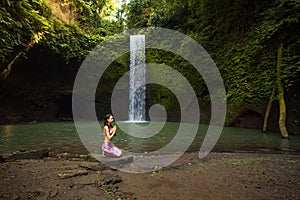 Young Caucasian woman sitting on the stone near the waterfall in Vajrasana or Diamond pose. Hands in namaste mudra. Tropical