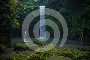 Young Caucasian woman sitting on the stone near the waterfall in Vajrasana or Diamond pose. Hands raised up in namaste mudra. Yoga