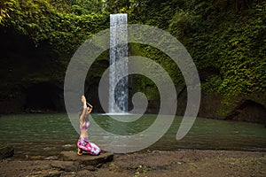 Young Caucasian woman sitting on the stone near the waterfall in Vajrasana or Diamond pose. Hands raised up in namaste mudra. Yoga