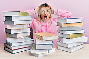 Young caucasian woman sitting on the table with books crazy and scared with hands on head, afraid and surprised of shock with open