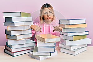 Young caucasian woman sitting on the table with books pointing down looking sad and upset, indicating direction with fingers,