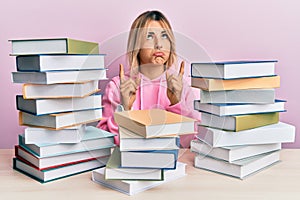 Young caucasian woman sitting on the table with books pointing up looking sad and upset, indicating direction with fingers,