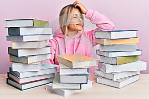 Young caucasian woman sitting on the table with books smiling confident touching hair with hand up gesture, posing attractive and