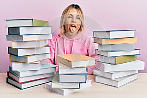 Young caucasian woman sitting on the table with books sticking tongue out happy with funny expression