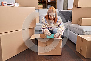 Young caucasian woman smiling confident unpacking books cardboard box at new home