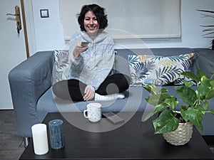 Young caucasian woman smiling and using the TV remote controller in pajamas, sitting on the sofa with a cup of coffee