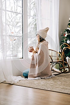 Young caucasian woman standing near window and looking outside in the morning, drinking tea and enjoying winter time