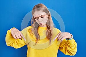 Young caucasian woman standing over blue background pointing down looking sad and upset, indicating direction with fingers,