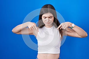 Young caucasian woman standing over blue background pointing down looking sad and upset, indicating direction with fingers,