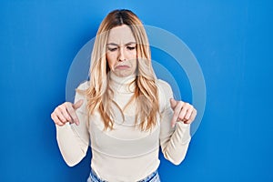 Young caucasian woman standing over blue background pointing down looking sad and upset, indicating direction with fingers,