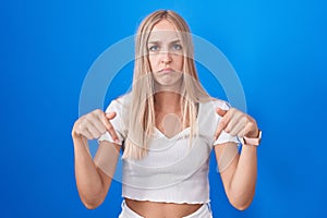 Young caucasian woman standing over blue background pointing down looking sad and upset, indicating direction with fingers,