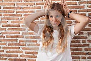 Young caucasian woman standing over bricks wall doing bunny ears gesture with hands palms looking cynical and skeptical