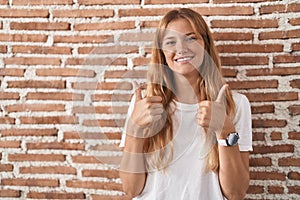 Young caucasian woman standing over bricks wall success sign doing positive gesture with hand, thumbs up smiling and happy