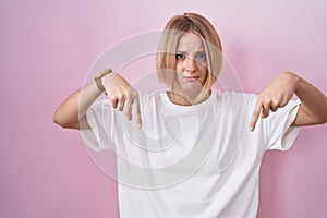 Young caucasian woman standing over pink background pointing down looking sad and upset, indicating direction with fingers,