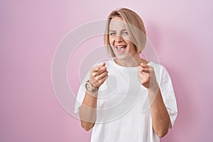 Young caucasian woman standing over pink background pointing fingers to camera with happy and funny face