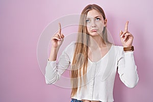 Young caucasian woman standing over pink background pointing up looking sad and upset, indicating direction with fingers, unhappy