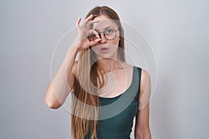 Young caucasian woman standing over white background doing ok gesture shocked with surprised face, eye looking through fingers