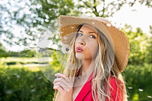 Young caucasian woman in a straw hat blowing on a dandelion