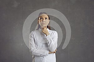 Young caucasian woman thinking and touching chin looking up stand over grey wall