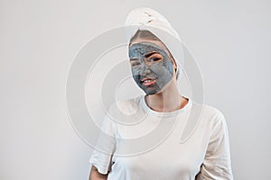 young caucasian woman in towel on head with black clay or mud facial mask isolated on white