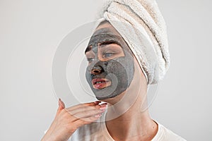 young caucasian woman in towel on head with black clay or mud facial mask isolated on white