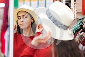 A young Caucasian woman tries on a straw hat for a vacation, and looks in the mirror. Close up. The concept of buying clothes