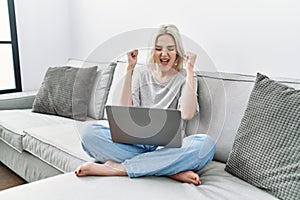 Young caucasian woman using laptop at home sitting on the sofa excited for success with arms raised and eyes closed celebrating