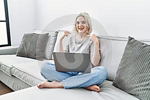 Young caucasian woman using laptop at home sitting on the sofa looking confident with smile on face, pointing oneself with fingers