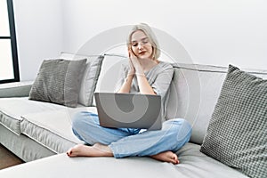 Young caucasian woman using laptop at home sitting on the sofa sleeping tired dreaming and posing with hands together while