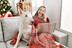 Young caucasian woman using laptop and talking on the smartphone sitting with dog by christmas tree at home