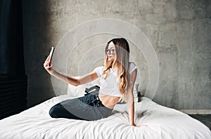 Young caucasian woman using mobile phone on bed at home, making selfie photo