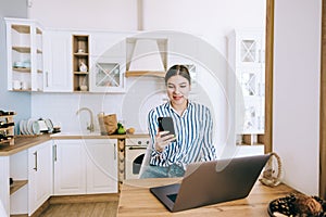 Young caucasian woman using mobile phone and laptop computer working at home, sitting on the kitchen at home