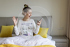 Young caucasian woman using mobile phone and wireless headphones, resting, sitting on bed at home and listening podcast