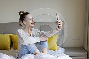 Young caucasian woman using mobile phone and wireless headphones, resting, sitting on bed at home and listening podcast