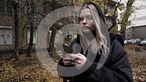 Young caucasian woman using phone in the city sleeping area in front of residential house in autumn. Action. Blond