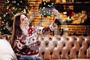Young caucasian woman using smart phone video call talking with friend or boyfriend. Christmas tree decorated with