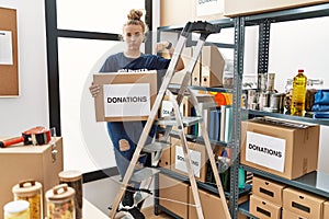 Young caucasian woman volunteer holding donations box pointing down looking sad and upset, indicating direction with fingers,